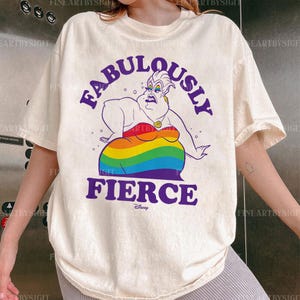 Peut inclure: T-shirt cr&egrave;me avec une illustration de dessin anim&eacute; d'Ursula de La Petite Sir&egrave;ne de Disney. Le personnage est repr&eacute;sent&eacute; avec une jupe aux couleurs de l'arc-en-ciel et les mots "FABULOUSLY FIERCE" en violet. Le t-shirt a des manches courtes.