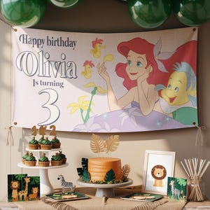 May include: A birthday party scene with a banner featuring Ariel from The Little Mermaid. The banner reads "Happy birthday Olivia Is turning 3." The scene includes a cake, cupcakes, and safari-themed decorations.