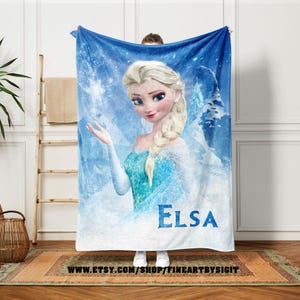 Puede incluir: Una manta polar azul con un retrato de Elsa de la pel&iacute;cula Frozen. Elsa est&aacute; representada con cabello rubio, un vestido azul y un copo de nieve. La palabra "Elsa" est&aacute; impresa en azul. La manta tiene un tema invernal.