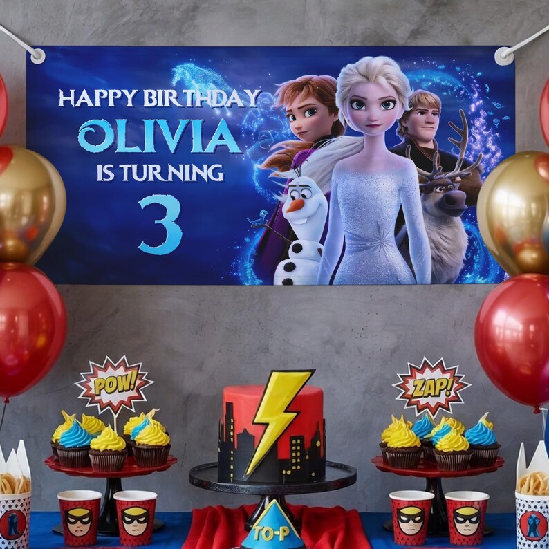 Puede incluir: Una escena de fiesta de cumplea&ntilde;os con una pancarta que dice "Feliz Cumplea&ntilde;os Olivia cumple 3 a&ntilde;os". La pancarta presenta personajes de la pel&iacute;cula Frozen. Una tarta roja y amarilla con un dise&ntilde;o de rayo se encuentra sobre un soporte negro. Tambi&eacute;n se ven cupcakes y art&iacute;culos de fiesta.