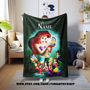May include: A dark green fleece blanket featuring a colorful illustration of Ariel from The Little Mermaid. The image includes Ariel, Flounder, Sebastian, and other characters. The blanket has the word "Name" at the top and the Etsy shop URL at the bottom.