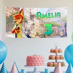 Pu&ograve; includere: Banner di compleanno con i personaggi di Zootropolis e la scritta "AMELIA COMPIE 3". La scena include una torta rosa, cupcakes e decorazioni per la festa.