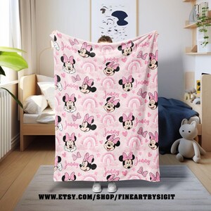 K&ouml;nnte beinhalten: Eine rosa Decke mit einem sich wiederholenden Muster von Minnie Mouse, Regenbogen, Herzen und Schleifen. Der Name "Jenny" ist in Schreibschrift im gesamten Design geschrieben. Die Decke wird in einem Kinderzimmer gezeigt.