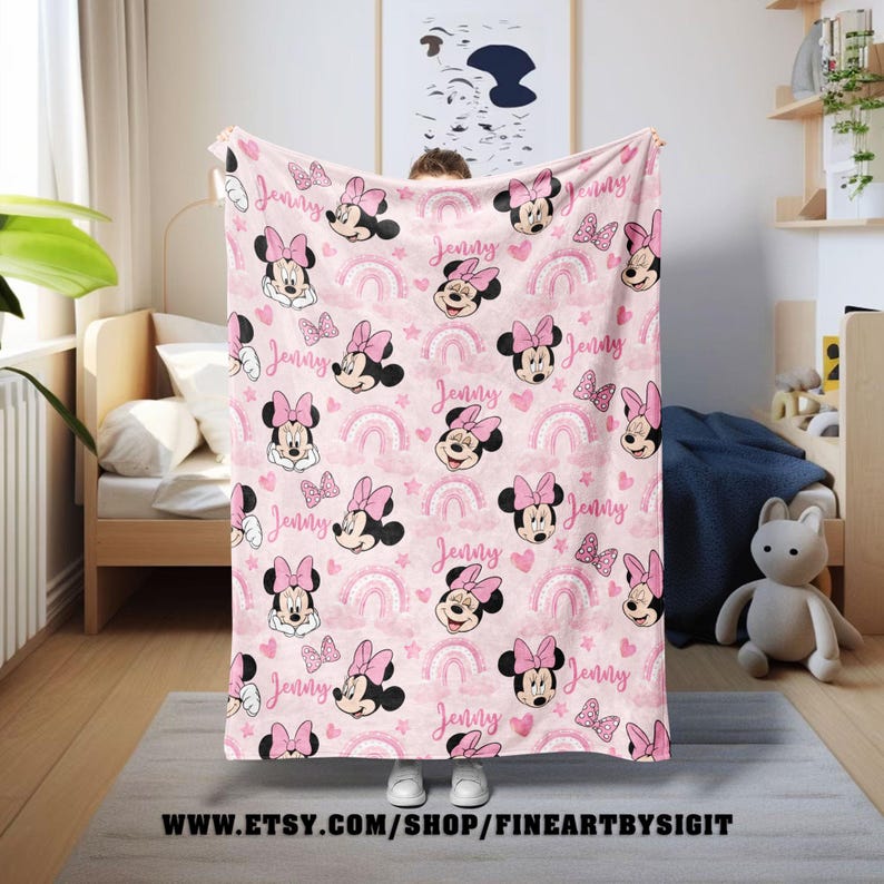 K&ouml;nnte beinhalten: Eine rosa Decke mit einem sich wiederholenden Muster von Minnie Mouse, Regenbogen, Herzen und Schleifen. Der Name "Jenny" ist in Schreibschrift im gesamten Design geschrieben. Die Decke wird in einem Kinderzimmer gezeigt.