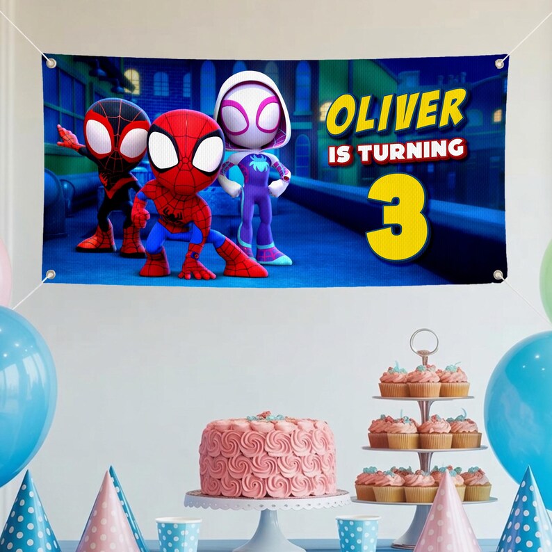 K&ouml;nnte beinhalten: Blaues Banner mit Spider-Man-Cartoonfiguren und dem Text "Oliver wird 3". Partydekorationen umfassen einen rosa Kuchen, Cupcakes, Luftballons und Partyh&uuml;te. Geeignet f&uuml;r eine Geburtstagsfeier.