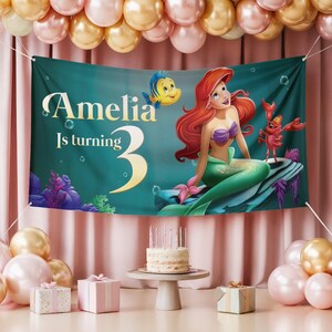 May include: A teal banner with the text "Amelia Is turning 3" and a mermaid illustration. The mermaid has red hair, a purple top, and a green tail. A yellow fish and red crab are also featured. The banner is part of a birthday party setup.
