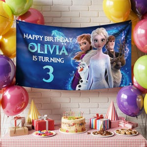 Puede incluir: Una pancarta de cumplea&ntilde;os con el texto "Happy Birthday Olivia is turning 3" con personajes de la pel&iacute;cula Frozen. La pancarta se muestra con globos, regalos, una tarta y gorros de fiesta.