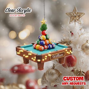 May include: A clear acrylic Christmas ornament shaped like a miniature pool table with a pyramid of billiard balls. The pool table has a teal felt surface, wooden frame, and string lights. The ornament has the text "Clear Acrylic" and "William".