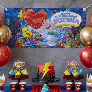 K&ouml;nnte beinhalten: Eine Geburtstagsparty-Szene mit einem Banner mit Arielle, der Meerjungfrau. Ein roter und schwarzer Kuchen mit einem gelben Blitz sitzt auf einem St&auml;nder. Cupcakes, Luftballons und Tassen im Superhelden-Stil vervollst&auml;ndigen die festliche Dekoration.