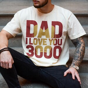 May include: A cream colored t-shirt with the text "DAD I LOVE YOU 3000" in red and gold. The text is stylized to look like the Iron Man character.