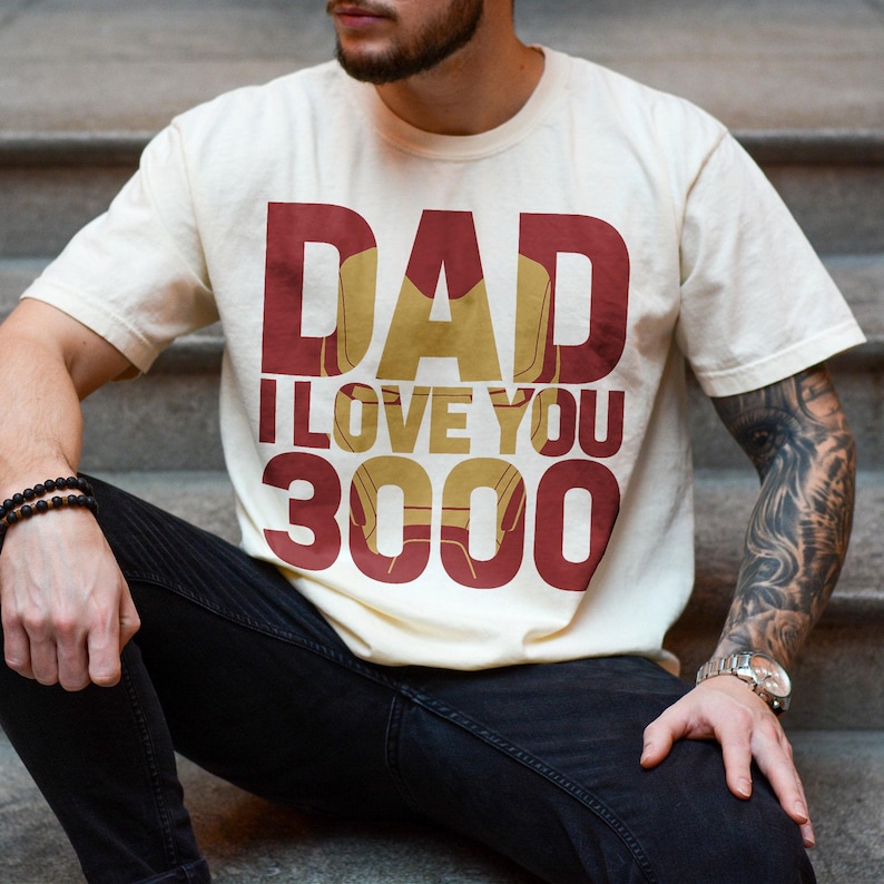 May include: A cream colored t-shirt with the text "DAD I LOVE YOU 3000" in red and gold. The text is stylized to look like the Iron Man character.