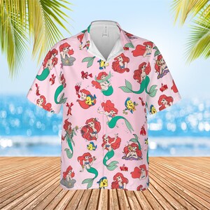 May include: A light pink short-sleeve button-down shirt featuring an all-over print of Ariel the mermaid and Flounder the fish in various poses. The shirt has a classic collar, a white inner neckline, and a repeating pattern of aquatic characters.