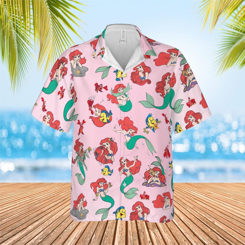 May include: A light pink short-sleeve button-down shirt featuring an all-over print of Ariel the mermaid and Flounder the fish in various poses. The shirt has a classic collar, a white inner neckline, and a repeating pattern of aquatic characters.