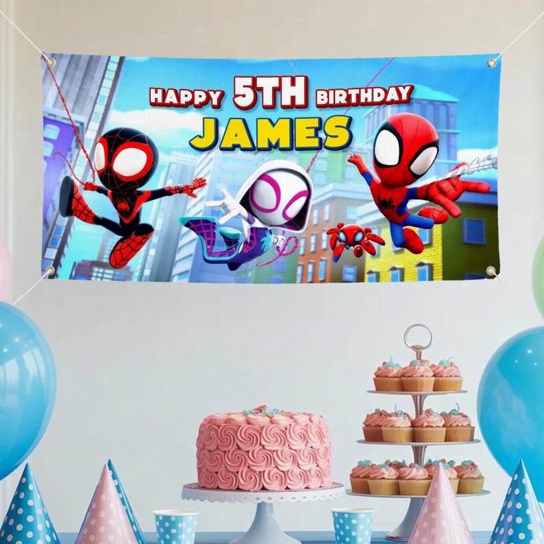 May include: A banner with the text "HAPPY 5TH BIRTHDAY JAMES" and images of Spider-Man characters. The banner is blue with a cityscape background. A pink cake and cupcakes are on a table with balloons.