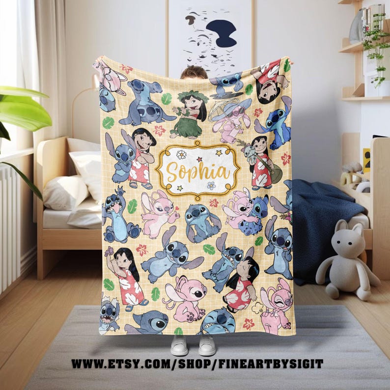 Puede incluir: Una manta beige con personajes de dibujos animados en varias poses. La manta tiene un patr&oacute;n repetido de personajes azules y rosas, con una etiqueta central con marco dorado que dice "Sophia". La manta se muestra en interiores.