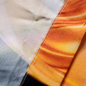May include: Close-up of fabric with a vibrant orange and yellow pattern, reminiscent of a sunset or fire. The fabric is divided by a seam with white stitching, creating a visual contrast. The texture of the fabric is visible.