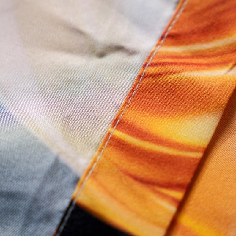 May include: Close-up of fabric with a vibrant orange and yellow pattern, reminiscent of a sunset or fire. The fabric is divided by a seam with white stitching, creating a visual contrast. The texture of the fabric is visible.