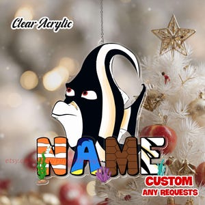 May include: Clear acrylic ornament featuring a cartoon fish design with the word "NAME" in colorful letters. The ornament has a black, white, and yellow fish design. The text "Clear Acrylic" and "CUSTOM ANY REQUESTS" are also visible.
