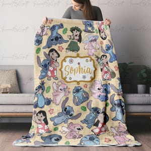Puede incluir: Una manta beige con personajes de dibujos animados en varias poses. La manta tiene un patr&oacute;n repetido de Stitch y Lilo, con el nombre "Sophia" en un marco decorativo. La manta se muestra en un sof&aacute;.