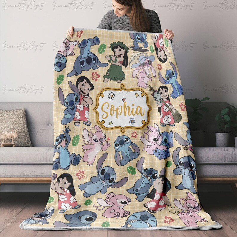 Puede incluir: Una manta beige con personajes de dibujos animados en varias poses. La manta tiene un patr&oacute;n repetido de Stitch y Lilo, con el nombre "Sophia" en un marco decorativo. La manta se muestra en un sof&aacute;.