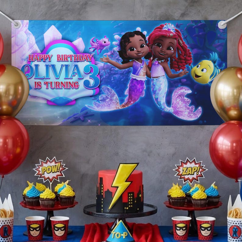 Peut inclure: D&eacute;coration de f&ecirc;te d'anniversaire avec une banni&egrave;re repr&eacute;sentant des sir&egrave;nes et le texte "HAPPY BIRTHDAY OLIVIA 3 IS TURNING." La table pr&eacute;sente un g&acirc;teau avec un &eacute;clair, des cupcakes et des gobelets et d&eacute;corations sur le th&egrave;me des super-h&eacute;ros. Des ballons rouges et dor&eacute;s sont &eacute;galement pr&eacute;sents.