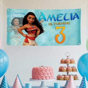 K&ouml;nnte beinhalten: Blaues Banner mit Moana-Illustration und dem Text "AMELIA IS TURNING 3". Geeignet f&uuml;r eine Geburtstagsfeier. Ein rosa Kuchen und Cupcakes sind ebenfalls zu sehen, zusammen mit Luftballons und Partyh&uuml;ten.