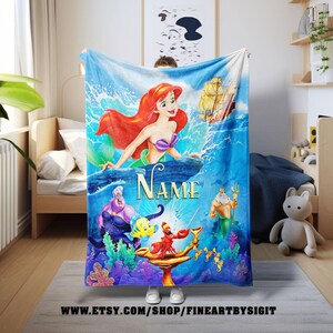 May include: A vibrant blue blanket featuring characters from The Little Mermaid. Ariel, Ursula, and King Triton are depicted in a colorful underwater scene. The word "NAME" is displayed prominently. The blanket has a nautical theme.