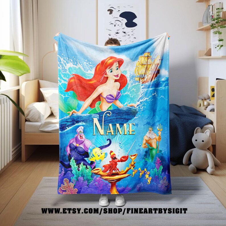 May include: A vibrant blue blanket featuring characters from The Little Mermaid. Ariel, Ursula, and King Triton are depicted in a colorful underwater scene. The word "NAME" is displayed prominently. The blanket has a nautical theme.