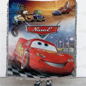 May include: A woven throw blanket featuring characters from the animated movie "Cars." The design includes Lightning McQueen, Mater, and other vehicles against a backdrop of a race track and desert landscape. The blanket has a fringed edge.