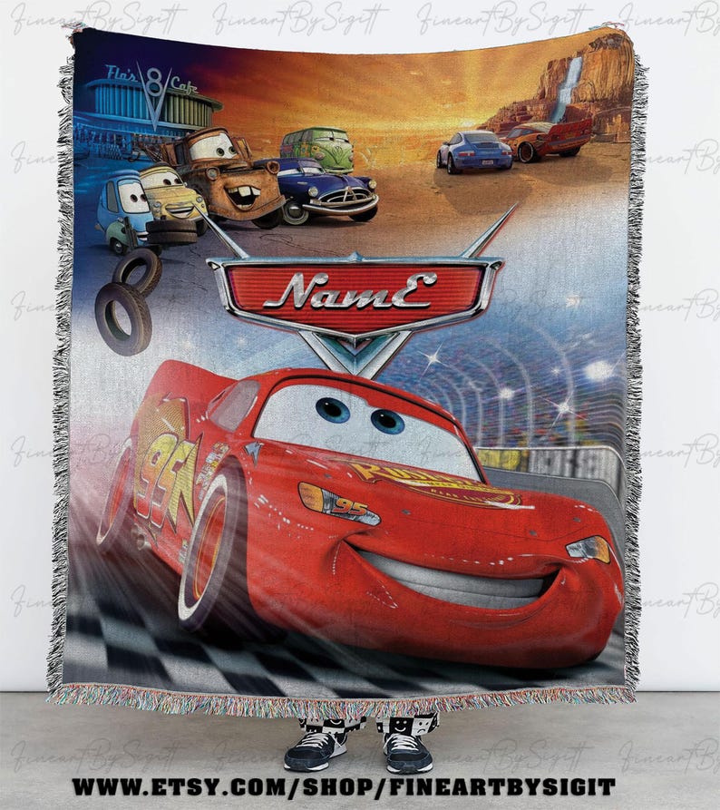 May include: A woven throw blanket featuring characters from the animated movie "Cars." The design includes Lightning McQueen, Mater, and other vehicles against a backdrop of a race track and desert landscape. The blanket has a fringed edge.