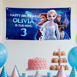 Puede incluir: Una pancarta azul con el texto "HAPPY BIRTHDAY OLIVIA IS TURNING 3" y personajes de la pel&iacute;cula Frozen. La pancarta presenta a Elsa, Anna, Kristoff, Olaf y Sven. Una tarta rosa y cupcakes sobre una mesa con globos azules y rosas.