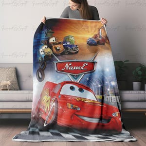 May include: A plush blanket featuring characters from the animated movie Cars. The design includes Lightning McQueen, Mater, and other vehicles against a backdrop of a race track and a sunset. The blanket has a checkered pattern.