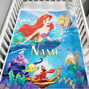 May include: A baby blanket featuring a colorful illustration of Ariel from The Little Mermaid. The blanket has a blue background with images of Ariel, Ursula, Flounder, Sebastian, King Triton, and a ship. The word "NAME" is printed in gold.