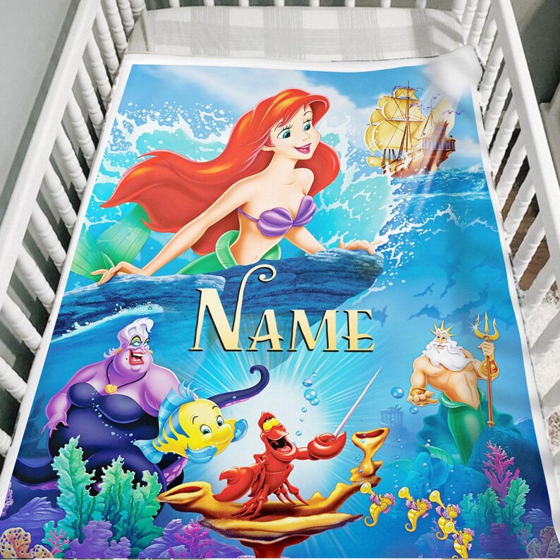 May include: A baby blanket featuring a colorful illustration of Ariel from The Little Mermaid. The blanket has a blue background with images of Ariel, Ursula, Flounder, Sebastian, King Triton, and a ship. The word "NAME" is printed in gold.