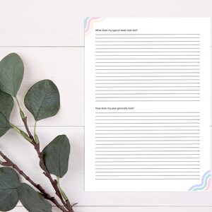 Pode incluir: Uma folha de trabalho imprim&iacute;vel com papel pautado e o texto "Como &eacute; a minha semana t&iacute;pica?" e "Como &eacute; o meu ano em geral?" A folha de trabalho est&aacute; sobre uma superf&iacute;cie de madeira branca com um ramo de folhas de eucalipto e uma caneta preta.