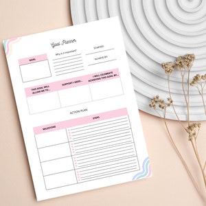 Pode incluir: Um planejador de objetivos imprim&iacute;vel com detalhes rosa e azul. O planejador inclui se&ccedil;&otilde;es para defini&ccedil;&atilde;o de objetivos, planos de a&ccedil;&atilde;o, marcos e etapas. O texto no planejador diz "Goal Planner".