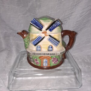 May include: A ceramic teapot shaped like a windmill with a green roof, blue and white blades, and brown handle. The teapot has a white base with brown trim and a green and brown floral design.