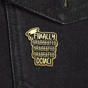 May include: Gold-coloured pin with the text "FINALLY GRAGEATED GRAGEATED GRAGEATED DONE!" and a graduation cap icon.