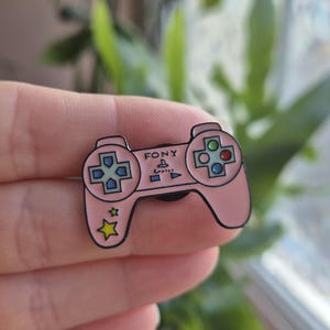 May include: A pink enamel pin in the shape of a retro video game controller. The pin has a black outline, colourful buttons, and the word "FONY" on the front. Two yellow stars are on the bottom left.