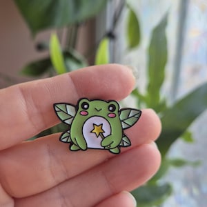 May include: A green and white enamel pin depicting a cartoon frog with fairy wings. The frog has a yellow star on its belly and pink cheeks. The pin is outlined in black.