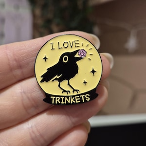 May include: A yellow enamel pin featuring a black crow with a purple gem, and the text "I LOVE TRINKETS". The pin is round with four small stars around the crow, and is a decorative item.