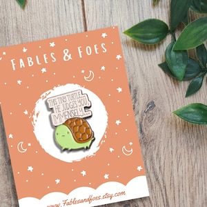 May include: A peach-coloured card with white stars and crescent moons printed on it. The card features a green and brown enamel pin of a turtle with a speech bubble that says "This tiny turtle... he judges you immensely". The card also has the text "Fables & Foes" and "www.Fablesandfoes.etsy.com" printed on it.
