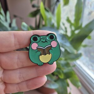May include: A green enamel pin depicting a cartoon frog reading a book. The frog has black eyes, pink cheeks, and a yellow belly. The pin is held in a hand against a backdrop of green leaves.