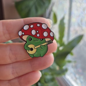 May include: A green frog playing a yellow banjo under a red mushroom cap with white spots. This enamel pin features a whimsical design, ideal for adding a touch of nature-inspired charm to clothing or accessories.