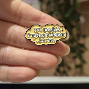 May include: A yellow enamel pin with the phrase "it's me, hi! I'm the problem it's me" in a retro font. The pin has a black outline and is shaped like a cloud with sparkling accents.