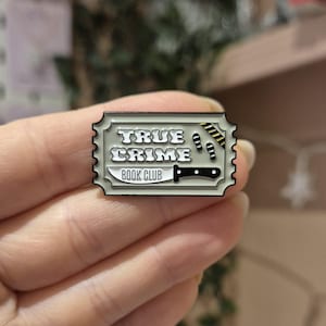 Könnte beinhalten: Rechteckiger Emaille-Pin mit Ticket-Design. Der Pin zeigt den Text "TRUE CRIME BOOK CLUB" in weißer, stilisierter Schrift. Ein Messer, Fußabdrücke und Absperrband sind ebenfalls abgebildet.