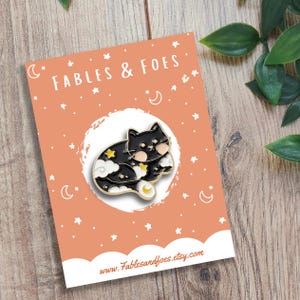 May include: A black cat enamel pin with a celestial design featuring stars, clouds, and a crescent moon. The pin is on a peach-coloured card with the text "FABLES & FOES" and the website address www.Fablesandfoes.etsy.com.