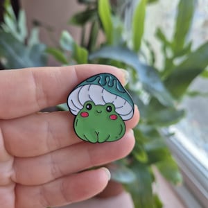 May include: A green frog with red cheeks and black eyes under a teal mushroom cap. The frog pin is a cute, cartoon-style design. The pin is held in a hand in front of a blurred background of green plants.