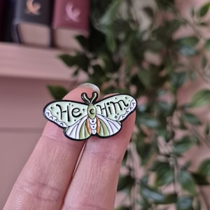 May include: A moth-shaped enamel pin with the words "He Him" in white script. The pin has a black outline, a light green body, and orange and white wing details. The pin is held in a hand.