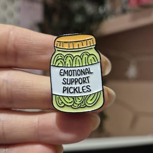 May include: A pin badge depicting a jar of pickles. The jar has a yellow lid and a white label with the text "EMOTIONAL SUPPORT PICKLES". The pickles inside are green.